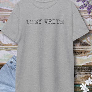 May include: A heather gray t-shirt with the text "THEY WRITE" in a dark gray font. The shirt is hanging on a wooden hanger. The background is a wooden wall with a pair of blue jeans.