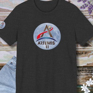 May include: Dark gray t-shirt featuring the Artemis II logo. The logo includes a stylized A, a red and blue arc, and the text "ARTEMIS II" against a moon background. A smaller logo is on the left sleeve.