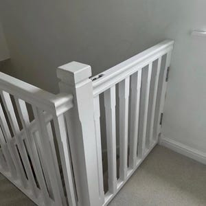 May include: A white painted wooden staircase balustrade with vertical spindles. The balustrade is positioned at a corner, with a carpeted floor visible below. The design is simple and classic.