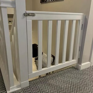 May include: A white wooden stair gate with vertical bars and a latch. A dog with black and white markings is behind the gate. The carpet has a grey and white zigzag pattern. The gate is designed to prevent access to the stairs.