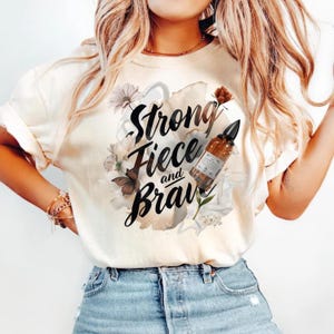 May include: Cream-colored t-shirt with the words "Strong, Fierce, and Brave" in black script, surrounded by floral and butterfly graphics. A brown bottle with a black cap is also featured.