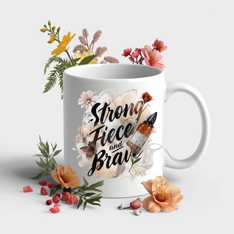 May include: White ceramic mug with the words "Strong, Fierce, and Brave" in black script, surrounded by floral and butterfly illustrations. A small brown bottle with a black dropper is also featured on the mug. The mug is decorated with peach and yellow flowers.