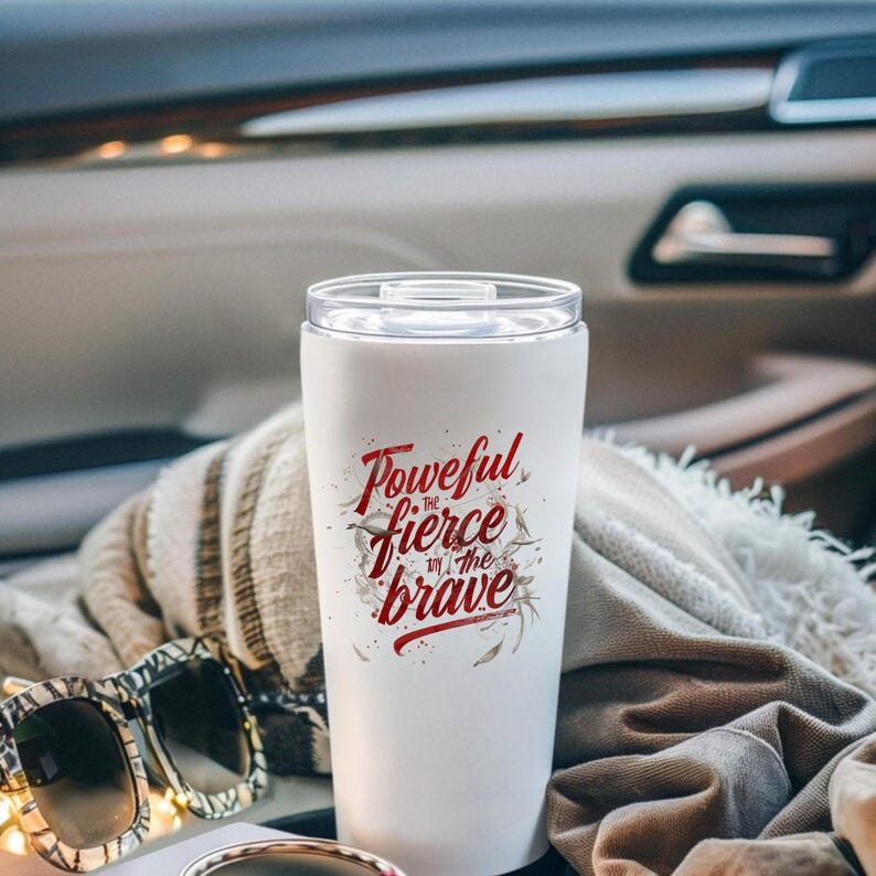 May include: White travel tumbler with a clear lid. The tumbler features the text "Powerful, fierce, in the brave" in red script. Sunglasses and a patterned blanket are in the foreground.