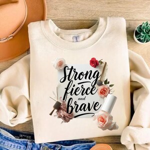 May include: Cream-colored sweatshirt with the words "Strong, fierce, and brave" in black script, surrounded by floral accents. The design includes pink and red roses, and nail polish. The sweatshirt is displayed on a wooden surface.