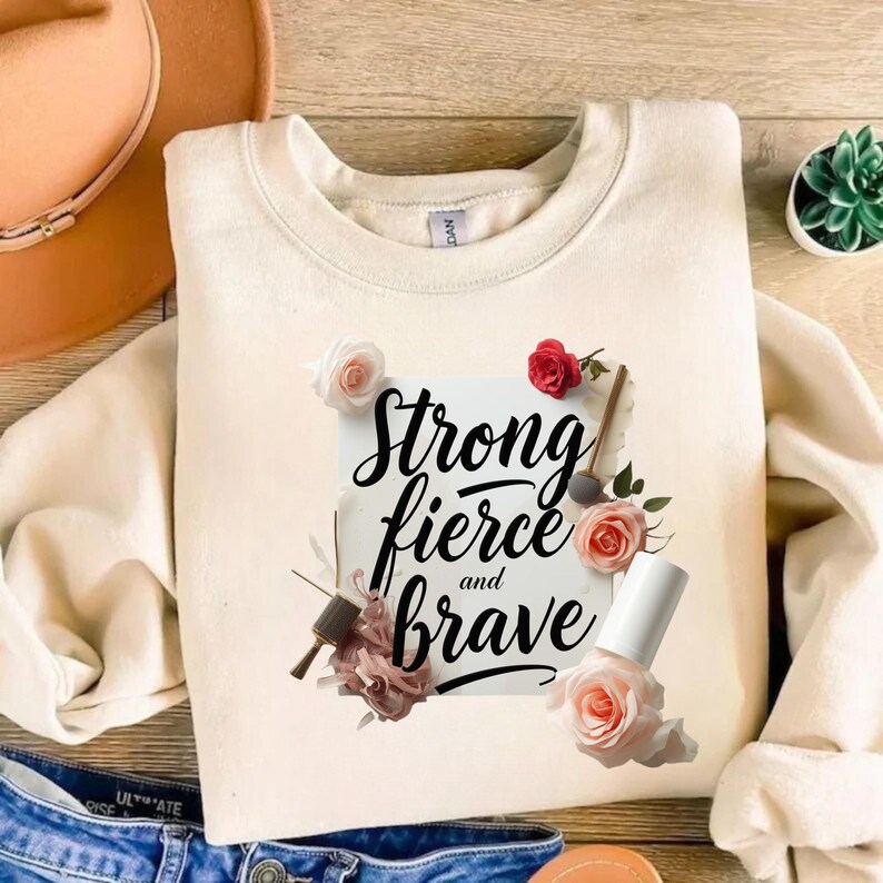 May include: Cream-colored sweatshirt with the words "Strong, fierce, and brave" in black script, surrounded by floral accents. The design includes pink and red roses, and nail polish. The sweatshirt is displayed on a wooden surface.