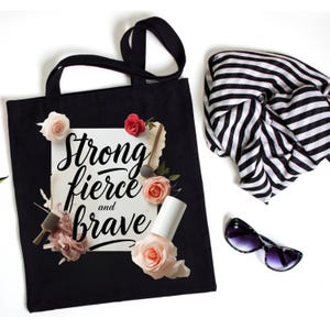 May include: Black tote bag with the words "Strong, fierce, and brave" in script. The bag is decorated with pink and red roses, a nail polish bottle, and a makeup brush. A black and white striped scarf and sunglasses are also in the image.