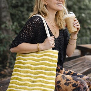 Puede incluir: Una bolsa de mano a rayas amarillas y blancas con el texto "Your Studio" es sostenida por una persona. La persona lleva una camiseta negra, gafas de sol y una diadema estampada. Sostiene una bebida con una pajita.