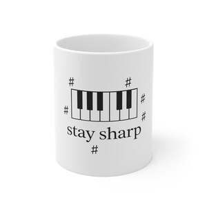 Puede incluir: Taza de cerámica blanca con un diseño de teclado de piano en blanco y negro y el texto "stay sharp" en negro. El diseño incluye símbolos musicales de sostenido. Un regalo temático musical.