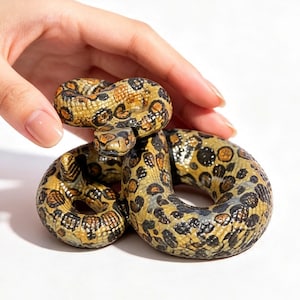 May include: A coiled snake figurine featuring a textured surface with a pattern of dark brown and black spots over a golden-yellow base. The snake is depicted in a resting position with its head raised slightly, showcasing detailed scales and eyes.