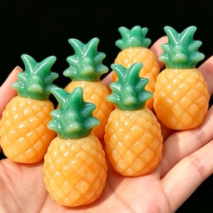 Op de afbeelding: Vijf kleine ananasvormige beeldjes gesneden uit steen. Elk stuk heeft een getextureerd, goudoranje lichaam met een gladde, doorschijnende groene bladeren top. De ananas-snijwerken zijn samen gerangschikt en vertonen een uniform ontwerp.