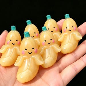 Könnte beinhalten: Sechs kleine, gelbe, bananenförmige Figuren mit lächelnden Gesichtern, rosa Wangen und blaugrünen Stielen. Jede Figur zeigt ein teilweise geschältes Bananendesign mit einer glatten, glänzenden Oberfläche, die in einer Gruppe angeordnet ist.