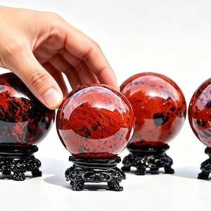May include: Four polished spherical stones with a deep red and black mottled pattern, each resting on a small, ornate black carved wooden stand. The spheres have a smooth, reflective surface and are arranged in a row against a plain background.