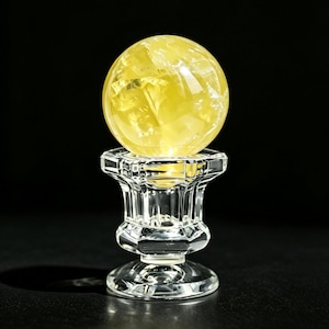 May include: A translucent yellow crystal sphere resting on a clear, faceted glass pedestal stand. The stand features a wide, octagonal top rim, a tapered neck, and a circular base. The text "SPHERE BASE, EXCLUDING THE CRYSTAL BALL" is visible.