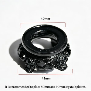Puede incluir: Un soporte de exhibición circular negro con detalles tallados ornamentados y tres patas. La abertura superior mide 40mm de diámetro y la base 45mm. El texto indica: It is recommended to place 50mm and 90mm crystal spheres.