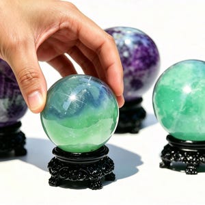 Può includere: Quattro sfere di cristallo nelle tonalità del verde, viola e blu, ciascuna su un supporto decorativo nero. Una mano tocca la sfera verde e blu. Le sfere sono illuminate, mostrando le loro qualità traslucide.