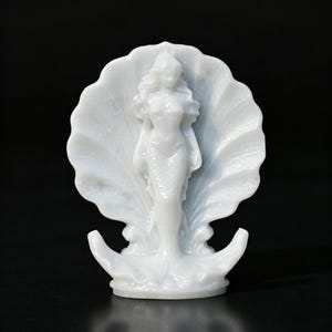 May include: A white figurine depicting a mermaid standing in the center of a large, open scallop shell. The mermaid has long, flowing hair and a defined tail, while the shell features prominent, radiating ridges. The entire piece is monochromatic.