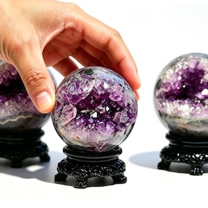 May include: Three spherical amethyst geodes, each featuring a polished outer surface and a deep purple crystalline interior, are displayed on ornate black stands. The spheres are arranged in a row, showcasing the natural violet crystal formations.