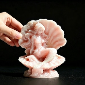 Könnte beinhalten: Eine hellrosa Wachsskulptur einer Figur mit langem, fließendem Haar, die in einer offenen Muschel sitzt. Die Skulptur hat eine glatte, halbtransparente Oberfläche und ist wie eine Muschelschale geformt, in der eine Figur mittig ruht.