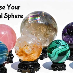 Op de afbeelding: Een verzameling van zeven kristallen bollen op zwarte standaards, waaronder roze, blauwe, doorzichtige, gele, groene en paarse varianten. De tekst "Choose Your Crystal Sphere" staat in zwarte letters linksboven tegen een witte achtergrond.