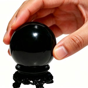 May include: A glossy black crystal ball, resting on a decorative black stand. The spherical object is held by a hand, suggesting its use for divination or display. The stand features ornate carvings.
