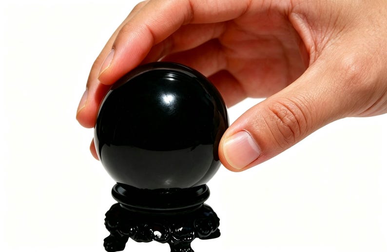 May include: A glossy black crystal ball, resting on a decorative black stand. The spherical object is held by a hand, suggesting its use for divination or display. The stand features ornate carvings.