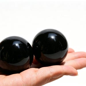 May include: Two polished, black obsidian spheres rest in an open palm against a white background. The spheres are smooth and reflective, with a glossy finish. The image is well-lit, highlighting the dark color and shape of the stones.