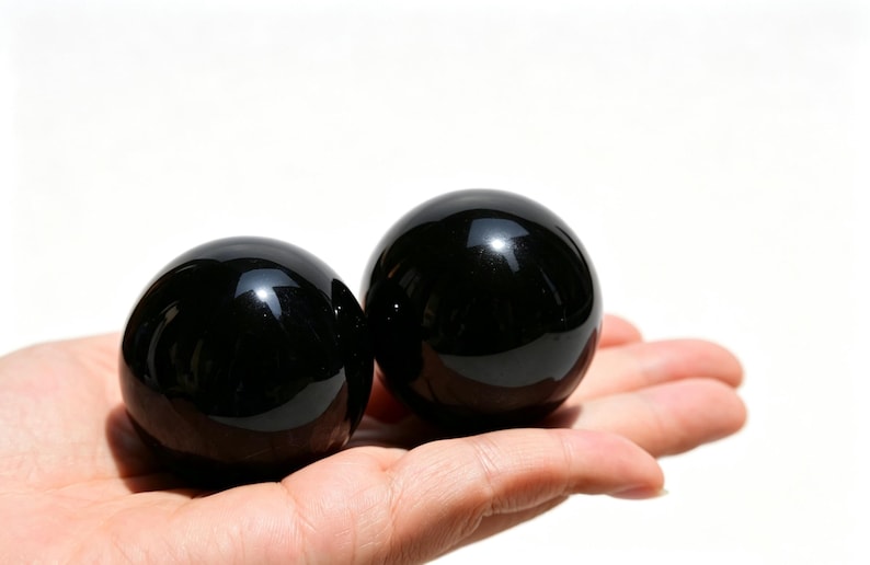 May include: Two polished, black obsidian spheres rest in an open palm against a white background. The spheres are smooth and reflective, with a glossy finish. The image is well-lit, highlighting the dark color and shape of the stones.