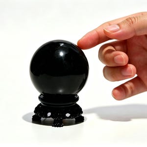 May include: A black crystal ball on a decorative black stand. The sphere is smooth and reflective, with a hand gently touching its surface. The stand has a detailed, ornate design. The background is white.