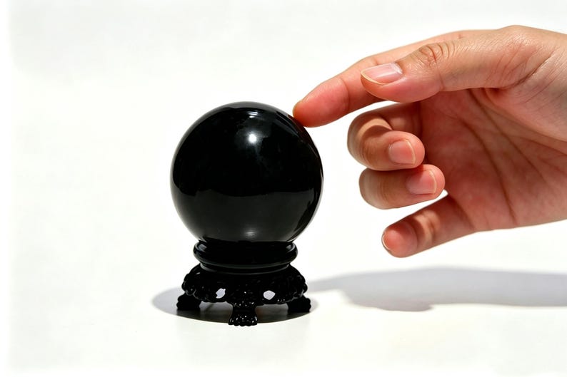 May include: A black crystal ball on a decorative black stand. The sphere is smooth and reflective, with a hand gently touching its surface. The stand has a detailed, ornate design. The background is white.