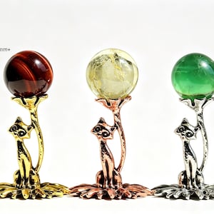 Puede incluir: Tres soportes decorativos de metal con forma de gato, cada uno sosteniendo una esfera de 30mm+. Los soportes tienen acabados en tono dorado, cobre y plata. Las esferas son de color rojo oscuro, amarillo pálido y verde. Cada gato se asienta sobre una base con forma de flor.