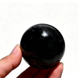 May include: A glossy, black sphere, held in a hand against a white background. The smooth surface reflects light, creating highlights. The object is a solid, round shape, likely a decorative item or a crystal ball.