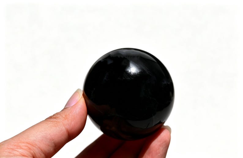 May include: A glossy, black sphere, held in a hand against a white background. The smooth surface reflects light, creating highlights. The object is a solid, round shape, likely a decorative item or a crystal ball.