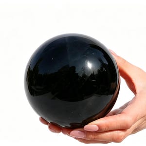 May include: A large, polished black sphere, held in a person's hands. The sphere reflects the surrounding environment, creating a mirrored effect. The object is a solid, dark color with a smooth, glossy surface.