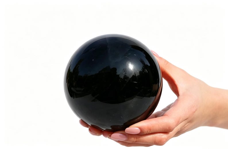 May include: A large, polished black sphere, held in a person's hands. The sphere reflects the surrounding environment, creating a mirrored effect. The object is a solid, dark color with a smooth, glossy surface.