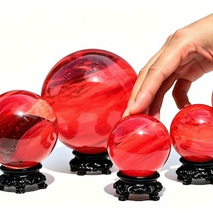 Op de afbeelding: Vier gladde, doorschijnende rode glazen bollen van verschillende groottes, elk rustend op een decoratieve zwart gesneden standaard. De bollen vertonen interne wervelende patronen van lichtere en donkerdere rode tinten.