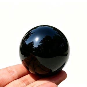 May include: A polished, black obsidian sphere. The surface reflects the surrounding environment, including trees and sky. The sphere is held in a hand against a white background. This decorative sphere is a unique home decor item.