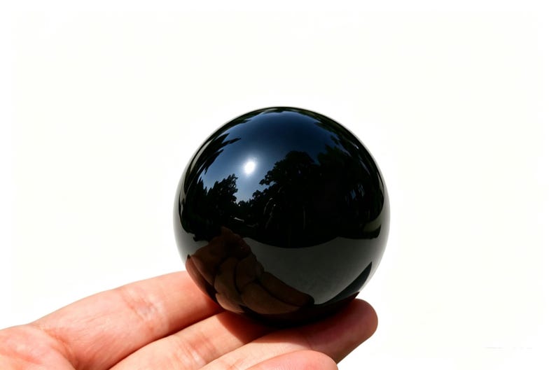 May include: A polished, black obsidian sphere. The surface reflects the surrounding environment, including trees and sky. The sphere is held in a hand against a white background. This decorative sphere is a unique home decor item.
