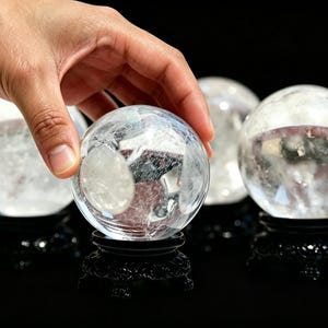 May include: A clear, spherical crystal ball with visible internal fractures and inclusions, resting on a decorative black carved stand. The sphere is transparent and smooth, reflecting light across its surface, set against a dark background.