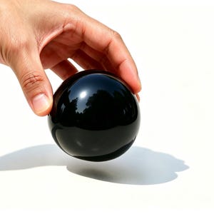 May include: A glossy, black sphere, reflecting light, is held by a hand against a white background. The smooth, dark surface of the ball contrasts with the bright backdrop, creating a striking visual.