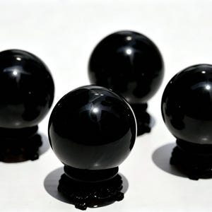 May include: Four polished black spheres, each resting on a dark, ornate base. The spheres have a glossy, reflective surface, and the bases cast shadows on the white background. The arrangement suggests a decorative or spiritual theme.