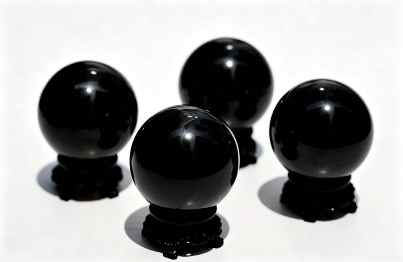 May include: Four polished black spheres, each resting on a dark, ornate base. The spheres have a glossy, reflective surface, and the bases cast shadows on the white background. The arrangement suggests a decorative or spiritual theme.
