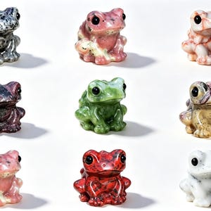 Peut inclure: Une collection de neuf petites figurines de grenouilles en pierre sculptée disposées en grille. Chaque grenouille présente une couleur et un motif uniques, notamment des tons gris, roses, verts, rouges et blancs, avec des surfaces polies.
