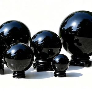May include: A collection of six polished black obsidian spheres of varying sizes, each resting on a black stand. The spheres reflect the surrounding environment, creating a glossy appearance. The spheres are arranged on a white surface.