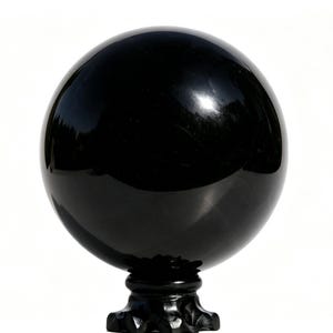 May include: A glossy, black sphere, possibly a decorative orb or gazing ball, sits atop a black, ornate pedestal. The sphere reflects the surrounding environment, creating a mirrored effect. The background is plain white.