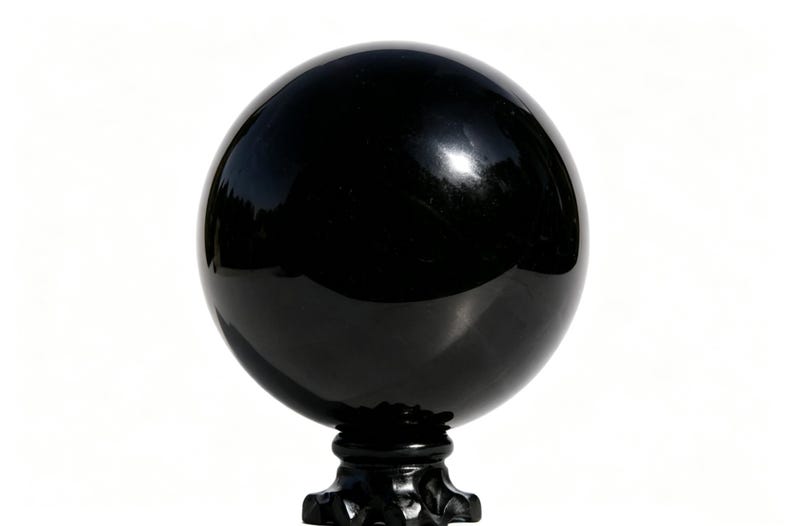 May include: A glossy, black sphere, possibly a decorative orb or gazing ball, sits atop a black, ornate pedestal. The sphere reflects the surrounding environment, creating a mirrored effect. The background is plain white.
