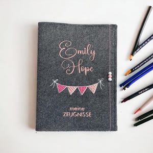 Könnte beinhalten: Grauer Filz-Notizbuchumschlag mit dem Namen "Emily Hope" in rosa Schrift gestickt. Unter dem Namen befindet sich ein Wimpelbanner in Rosa und Weiß. Der deutsche Text "meine ZEUGNISSE" ist unten gestickt. Eine rosa Schnur mit Perlen verläuft an der Seite.