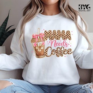 May include: White sweatshirt with the phrase "MAMA Needs Coffee" in a mix of brown checkered and pink script lettering. A graphic of an iced coffee with a pink bow and straw is on the left. The sweatshirt is a casual, everyday wear item.