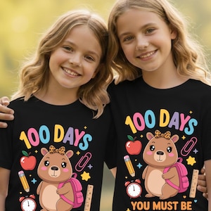 May include: Two girls wearing black t-shirts with a colorful graphic. The shirts feature a cartoon bear with a backpack, apple, pencil, and the text "100 DAYS YOU MUST BE CAPY."