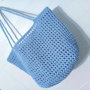 May include: A light blue, open-weave crochet tote bag. The bag has long, thin straps and a rounded bottom. The crochet pattern creates a textured appearance. The bag is hanging against a white background.