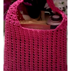 May include: A magenta crochet tote bag with a rectangular body and a handle. The bag has an open weave design and is filled with items. The background features purple flowers.